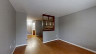 Located at 2407 W Vine St in Kissimmee, Florida, this single-floor unit offers a practical and efficiently designed layout comprising a living room and a full bathroom. The living area is finished with warm-toned hardwood-style flooring that extends throughout, complementing the neutral gray walls and white baseboards for a clean, cohesive look. A standout architectural feature is the mid-century-inspired open wood room divider, constructed with a rich dark wood frame, which adds character and visually segments the space without sacrificing openness. Natural light enters through a large floor-to-ceiling window with blinds adjacent to the front entry door, brightening the interior and offering a view of the surrounding greenery. A wall-mounted HVAC unit provides climate control. The space also includes a closet area with built-in shelving, visible from the main living zone. The bathroom is equipped with a full-size soaking tub and shower combination set within a clean white surround, along with a vanity featuring warm wood cabinetry and a wall mirror. The textured ceilings and consistent gray wall palette throughout give the unit a uniform, no-frills character well-suited for practical everyday living.