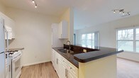 This single-floor space showcases a well-designed open-concept layout, where the kitchen flows seamlessly into the living area, creating a cohesive and functional environment. The kitchen is equipped with crisp white cabinetry featuring sleek bar-pull hardware, providing ample storage along both the upper and lower sections. Dark granite countertops offer a striking contrast against the white cabinets, running along the perimeter and extending into a peninsula that serves as a natural divider between the kitchen and the adjacent living space. The peninsula also provides additional prep area and casual seating potential. A stainless steel double-basin sink is positioned along the counter, accompanied by a modern gooseneck faucet. Track lighting overhead ensures the kitchen is well-lit, while light wood-look flooring runs throughout, contributing to a warm and cohesive aesthetic. A tile backsplash adds a refined detail behind the sink area. The living area beyond benefits from a large window that invites abundant natural light, enhancing the open and airy feel of the space. Neutral wall tones throughout keep the interior palette clean and versatile.
