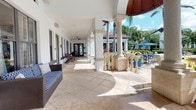 This single-story Advenir community clubhouse showcases a refined Mediterranean-inspired design that seamlessly blends indoor and outdoor living. The exterior is defined by striking cream-colored stone columns with ornate balustrades, set against crisp white stucco walls and a rich terracotta-toned domed entrance. Expansive travertine-tiled terraces wrap around the building, providing generous shaded walkways furnished with wicker seating — ideal for relaxing while taking in the surrounding scenery.

The poolside colonnade offers a graceful vantage point overlooking a sparkling swimming pool, framed by swaying palm trees and vibrant tropical plantings. Ceiling fans line the covered corridor, ensuring comfort throughout the warm seasons. Stone planters filled with bold tropical plants flank the entry steps, adding to the lush, resort-like ambiance.

Glass double doors bearing the Advenir branding provide an inviting entrance into the clubhouse, with reflections of the pool and columns visible through the glass — blurring the line between interior and exterior. The overall atmosphere is one of upscale leisure, characterized by abundant natural light, open-air vistas, and carefully curated architectural details that evoke a sense of a private resort retreat.