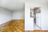 This thoughtfully laid-out 1-bedroom, 1-bathroom residence offers a comfortable and functional living environment across a single floor. Classic parquet hardwood flooring runs throughout the main living areas and bedroom, lending warmth and timeless character to the space.

The spacious living area is a standout feature, anchored by a wide bank of windows framing lush, tree-lined views. A radiator beneath the windows provides efficient heating, and the light-filled room offers generous space for entertaining and everyday living.

The kitchen is accessed through a dedicated doorway and features updated gray cabinetry, a striking granite countertop with subtle veining, a white tile backsplash, and white appliances. Light tile flooring completes the clean, modern aesthetic. An adjacent area accommodates laundry.

The bedroom is a well-proportioned retreat, benefiting from a window with greenery views and parquet flooring. Two separate doors provide access to both a closet and the hallway, offering practical storage and circulation.

The bathroom features a classic black-and-white tile design, with white field tiles accented by a bold black border, complemented by a full soaking tub with an overhead shower. A connecting hallway ties all rooms together for a cohesive and efficient floor plan.