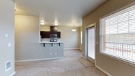 This single-floor space features an open-concept design that seamlessly connects the kitchen and living area. The kitchen is equipped with dark espresso cabinetry, stainless steel appliances including a microwave and range, and a raised breakfast bar with bar seating — an ideal setup for casual dining or entertaining. Flush-mount ceiling lighting illuminates the kitchen area, while a pendant light fixture adds a decorative touch above the bar.

The living area is finished with wall-to-wall carpeting in a neutral tone, contributing to a warm and comfortable atmosphere. Soft beige walls throughout maintain a clean, cohesive look. Natural light enters through multiple windows fitted with horizontal blinds, and a glass-paneled door — likely leading to a patio or exterior — further enhances the sense of brightness and connectivity to the outdoors. The open layout creates a spacious feel, with clear sightlines from the kitchen through to the living space. Overall, the unit presents a modern, move-in-ready aesthetic with functional design choices suited for everyday living.