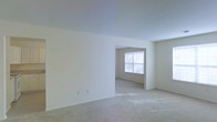 This single-floor space presents a clean, minimalist layout characterized by bright white walls and wall-to-wall neutral-toned carpeting throughout. The main living area is generously proportioned, offering an open feel that flows seamlessly into an adjacent room through a wide passageway, creating a sense of connected, flexible living space. The secondary room, visible through the opening, features a window fitted with horizontal blinds that allows natural light to filter in, casting soft illumination across the carpeted floor. The smooth, white ceilings maintain a consistent height throughout, contributing to the overall sense of openness. Electrical outlets are positioned along the baseboards, and the walls are free of ornamentation, presenting a blank canvas well-suited for personalization. The neutral color palette — from the white walls and ceilings to the beige carpeting — creates a cohesive, understated aesthetic throughout the space. Overall, the layout emphasizes practicality and openness, making it adaptable to a variety of furniture arrangements and functional uses.