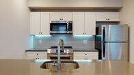 This single-floor residence showcases a thoughtfully designed open-concept layout, with warm hardwood-style flooring flowing throughout the main living areas. The spacious living room is lined with large floor-to-ceiling windows that frame urban views and fill the space with abundant natural light, complemented by recessed ceiling lighting.

The kitchen is a standout feature, offering white shaker-style cabinetry extending to the ceiling, stainless steel appliances including a range, over-the-range microwave, and refrigerator, alongside a striking herringbone-patterned tile backsplash with subtle under-cabinet LED accent lighting. A generously sized island with a sleek countertop and an undermount sink serves as a functional and visual centerpiece.

The bedrooms are carpeted in soft grey tones, each featuring windows with open city views, providing a comfortable and bright retreat. Storage is addressed with louvered sliding closet doors in the main living area.

The two bathrooms are finished with marble-style tile surrounds, matte black fixtures, white vanities with undermount sinks, and light tile flooring—one featuring a tub/shower combination and the other a glass-enclosed walk-in shower, both offering a clean, contemporary aesthetic.