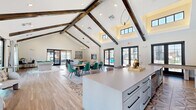 Located at 7211 Tutt Blvd in Colorado Springs, CO, this generously sized single-floor community clubhouse delivers an impressive blend of rustic and contemporary design. The defining architectural feature is the dramatic vaulted ceiling adorned with rich, dark wood beams that span the open-concept main area, creating a grand sense of height and volume throughout the space.

The flooring transitions elegantly between wide-plank wood-look planks and intricately patterned geometric tile, adding visual interest underfoot. A large central island with a sleek quartz countertop and bar seating serves as the heart of the space, complemented by a full-length counter run equipped with a coffee station and appliances. A striking floor-to-ceiling stone accent wall frames a mounted television, anchoring the lounge area where comfortable seating arrangements invite relaxation.

Natural light pours in through numerous black-framed French doors and clerestory windows, with views overlooking an outdoor pool and patio. A billiards table adds a recreational element, while a cozy windowed nook provides a quieter retreat with direct pool views. Recessed lighting and contemporary pendant fixtures complete the polished, welcoming ambiance of this well-appointed community gathering space.