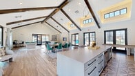 Located at 7211 Tutt Blvd in Colorado Springs, CO, this generously sized single-floor community clubhouse delivers an impressive blend of rustic and contemporary design. The defining architectural feature is the dramatic vaulted ceiling adorned with rich, dark wood beams that span the open-concept main area, creating a grand sense of height and volume throughout the space.

The flooring transitions elegantly between wide-plank wood-look planks and intricately patterned geometric tile, adding visual interest underfoot. A large central island with a sleek quartz countertop and bar seating serves as the heart of the space, complemented by a full-length counter run equipped with a coffee station and appliances. A striking floor-to-ceiling stone accent wall frames a mounted television, anchoring the lounge area where comfortable seating arrangements invite relaxation.

Natural light pours in through numerous black-framed French doors and clerestory windows, with views overlooking an outdoor pool and patio. A billiards table adds a recreational element, while a cozy windowed nook provides a quieter retreat with direct pool views. Recessed lighting and contemporary pendant fixtures complete the polished, welcoming ambiance of this well-appointed community gathering space.