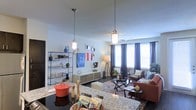 This single-floor space showcases an open-concept layout that seamlessly connects the kitchen and living areas. The kitchen features striking granite countertops and a dark cooktop surface, with two pendant lights—cylindrical glass fixtures with brass accents—hanging overhead to provide focused task lighting. A black metal open shelving unit along the kitchen wall displays decorative objects and accessories, adding an eclectic, curated touch to the space.

The living area is anchored by a low-profile media console with a rustic wood finish, flanked by wall art and a decorative mirror. A seating arrangement of a sofa and accent chairs surrounds a glass-top coffee table, all set atop a richly patterned area rug that adds warmth and texture to the dark flooring. A tripod floor lamp provides additional ambient lighting in the corner.

Large windows dressed with floor-length charcoal curtains line the far wall, allowing generous natural light to fill the space. A ceiling fan is centrally positioned above the living area. The overall ambiance is contemporary and thoughtfully styled, blending industrial, rustic, and modern design elements throughout.