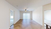 This well-designed single-floor residence offers a cohesive blend of modern finishes and functional living spaces. The open living area makes an immediate impression with its striking herringbone-patterned hardwood flooring, crisp white walls, and a ceiling fan with integrated lighting. A large sliding glass door fitted with vertical blinds fills the room with natural light and provides access to the outdoors. The kitchen is a standout feature, showcasing rich dark-toned cabinetry, light granite countertops, a sparkling mosaic tile backsplash, and a full suite of stainless steel appliances including a range, microwave, refrigerator, and dishwasher. A central island with pendant lighting adds both workspace and visual appeal. The herringbone flooring continues seamlessly from the living area into the kitchen, reinforcing a sense of continuity. The bedrooms feature plush neutral carpeting and generous windows fitted with roller shades, offering views of the surrounding urban rooftops and cityscape. Additional rooms with light hardwood flooring provide flexible living or workspace options. The bathroom is appointed with patterned tile flooring, a dark-toned vanity with stone countertop, and matte black hardware accents, adding a refined touch to the overall design.