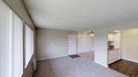 This single-floor space presents a practical and straightforward layout characterized by neutral, warm-beige walls and wall-to-wall carpeting that adds a sense of softness underfoot. The main living area is open and generously proportioned, offering ample room for a variety of furniture arrangements. A textured popcorn ceiling adds dimension overhead, while a ceiling fan with an integrated light fixture provides both air circulation and ambient lighting to the space.

A centrally positioned interior door serves as a focal point along the main wall, flanked by a row of built-in closet doors that extend across a significant portion of the wall. These built-ins offer considerable storage capacity, contributing to the room's overall functionality. White baseboard trim lines the perimeter of the room, providing a clean contrast against the beige walls. A window along one side of the room allows for natural light to enter, brightening the interior. A small heating unit is visible near the window, indicating climate control features within the space. Overall, the unit presents a clean, move-in ready environment with a neutral palette suited to a range of personal styles.