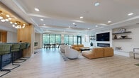 Located at 7510 N Oak Trafficway in Gladstone, Missouri, this single-floor community amenity space is a thoughtfully designed open-concept lounge that seamlessly blends comfort, function, and modern style. The layout is organized into several distinct yet interconnected zones, all unified by light wood-tone luxury vinyl plank flooring and tray ceilings with recessed lighting throughout.

The main lounge area serves as the centerpiece, featuring a large wall-mounted flat-screen television above a sleek linear fireplace, flanked by floating shelves. Plush tan leather sectional sofas and a curved accent chair create a welcoming gathering atmosphere. Adjacent to this is a dedicated dining area bathed in natural light from expansive floor-to-ceiling black-framed windows, which offer views of the surrounding tree-lined landscape and connect to an outdoor balcony.

A stylishly appointed kitchenette runs along one side of the space, equipped with dark navy cabinetry, open metal shelving with geometric tile backsplash, a stainless steel refrigerator, and a bar-height counter lined with olive green upholstered stools. Suspended rattan hanging chairs add a playful, resort-inspired touch. A quieter nook provides a cozy reading retreat with neutral armchairs and a warm wood open bookcase, offering a more intimate setting within the broader communal space.