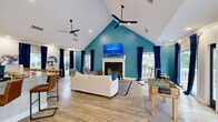 This single-floor clubhouse is a thoughtfully designed community gathering space that balances comfort, functionality, and lively character. The centerpiece is a spacious open-plan living area anchored by a dramatic vaulted ceiling that peaks over a bold teal accent wall. A white-mantled fireplace sits beneath a wall-mounted flat-screen TV, flanked by large windows dressed in rich navy curtains that frame outdoor views of a deck and surrounding greenery. Dual ceiling fans keep the airy space comfortable, while light wood-look flooring runs throughout, enhancing the sense of openness. Comfortable seating arrangements invite relaxed conversation, while a foosball table and additional recreation space cater to more active social gatherings. Glass doors open directly to an outdoor deck, seamlessly extending the living area. The kitchen is well-appointed with white shaker cabinetry, granite countertops, stainless steel appliances, and a breakfast bar with leather-upholstered stools — ideal for hosting events. The bathroom features striking black-and-white botanical wallpaper, a dark-finished vanity with a round mirror, and tile flooring, offering a stylish and functional retreat. Overall, this clubhouse delivers an inviting and versatile environment suited for a wide range of community activities.