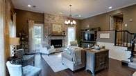 Located at 2225 Ashley River Road in Charleston, South Carolina, this single-story home blends classic Southern charm with comfortable, well-appointed interiors. The living area serves as the heart of the home, anchored by a striking floor-to-ceiling whitewashed brick fireplace that adds warmth and architectural character to the space. Rich, dark hardwood floors run throughout, providing a grounding contrast to the warm taupe walls and neutral furnishings. Natural light flows generously through multiple windows dressed with elegant floor-length curtains, offering views of the lush outdoor surroundings. A stylish chandelier hangs centrally, complementing the recessed lighting overhead to create a layered and inviting ambiance. The room is thoughtfully arranged with tufted seating, a plush area rug, and rustic accent pieces, including a distressed wood console that adds a touch of farmhouse-inspired character. Built-in shelving and a media center provide both functionality and display space along one wall. Crown molding and wainscoting details further elevate the interior's refined aesthetic. The overall design of this Charleston home reflects a timeless, transitional style that is both functional and visually inviting.