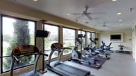 This fitness center is thoughtfully designed to provide an energizing workout environment. The space is lined with large windows along one wall, allowing abundant natural light to fill the room while offering views of the surrounding urban landscape and tree-lined scenery — an engaging backdrop for exercise.

The cardio area is well-stocked with a row of treadmills positioned parallel to the windows, complemented by elliptical trainers and stationary bikes arranged in a practical layout that maximizes floor space and ease of movement. Each cardio machine is equipped with a personal display monitor, allowing users to track workout metrics or follow entertainment.

Mounted flat-screen televisions on adjustable stands are strategically placed throughout the space, ensuring visibility from multiple workout stations. Overhead, ceiling fans with integrated lighting provide both air circulation and ambient illumination, contributing to a comfortable exercise environment.

The flooring includes both standard tile and rubberized mat sections, offering appropriate surfaces for different types of exercise. Recessed lighting in the textured ceiling supplements natural light, ensuring the space remains well-lit at all times. The overall atmosphere is functional, clean, and conducive to focused fitness activity.