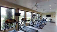 This fitness center is thoughtfully designed to provide an energizing workout environment. The space is lined with large windows along one wall, allowing abundant natural light to fill the room while offering views of the surrounding urban landscape and tree-lined scenery — an engaging backdrop for exercise.

The cardio area is well-stocked with a row of treadmills positioned parallel to the windows, complemented by elliptical trainers and stationary bikes arranged in a practical layout that maximizes floor space and ease of movement. Each cardio machine is equipped with a personal display monitor, allowing users to track workout metrics or follow entertainment.

Mounted flat-screen televisions on adjustable stands are strategically placed throughout the space, ensuring visibility from multiple workout stations. Overhead, ceiling fans with integrated lighting provide both air circulation and ambient illumination, contributing to a comfortable exercise environment.

The flooring includes both standard tile and rubberized mat sections, offering appropriate surfaces for different types of exercise. Recessed lighting in the textured ceiling supplements natural light, ensuring the space remains well-lit at all times. The overall atmosphere is functional, clean, and conducive to focused fitness activity.