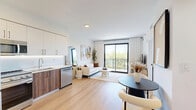 Located at 2479 NE 192nd St in Miami, FL, this well-appointed one-bedroom, one-bathroom residence offers a modern and cohesive design throughout its single-floor layout. Light-toned hardwood flooring flows seamlessly across the open-concept kitchen and living area, creating a bright and unified feel. The kitchen is outfitted with two-tone cabinetry—combining white upper cabinets with rich wood lower cabinets—complemented by a clean white quartz countertop, stainless steel appliances, and under-cabinet lighting. The living area is anchored by large sliding glass doors that open to a private balcony, flooding the space with natural light and offering views of lush greenery. The bedroom is a serene retreat featuring large windows with balcony access, delivering continued outdoor views and abundant daylight. A dedicated closet provides practical storage. The bathroom showcases a refined aesthetic with wood-panel tile surround on the soaking tub and shower, matte black fixtures, a wood-toned vanity, a backlit round mirror, and open floating shelves. A hallway connects the living spaces efficiently, completing this thoughtfully designed Miami residence.
