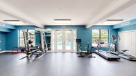 Located in Stuart, Florida, this fitness center offers a generously sized, single-floor space designed to support a comprehensive workout experience. The interior is defined by bold teal accent walls contrasted against crisp white ceilings and trim, creating an energetic yet clean aesthetic throughout. Durable rubber flooring covers the exercise areas, providing both safety and functionality underfoot.

The facility is thoughtfully organized to accommodate a wide range of fitness activities. A cardio zone features treadmills, stationary bikes, and an elliptical machine, while a strength training area includes a multi-station cable machine, a smith machine, a bench press setup, and a well-stocked dumbbell rack. Open floor space allows for stretching, functional training, and use of stability equipment such as medicine balls and plyometric boxes.

Natural light filters in through multiple windows and arched architectural details, with views of lush greenery and a resort-style pool just beyond the glass. French doors open directly to the pool area, seamlessly connecting indoor and outdoor spaces. Full-length mirrors along the walls enhance the sense of openness and allow users to monitor their form during workouts.

Flush-panel ceiling lights provide consistent, even illumination throughout, complementing the natural light. The space is served by a central HVAC system, ensuring year-round comfort for all users.