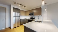 This well-designed one-bedroom, one-bathroom unit offers a thoughtful layout across a single floor. The open-concept kitchen and living area serves as the heart of the space, featuring light wood-style flooring that extends throughout. The kitchen is outfitted with a full suite of stainless steel appliances, including a refrigerator, electric range, built-in microwave, and dishwasher. Two-toned cabinetry — combining warm wood uppers with sleek grey lowers — pairs with white quartz countertops and a classic white tile backsplash. A peninsula with pendant lighting provides additional prep space and casual dining. The living area is bright and open, benefiting from floor-to-ceiling windows and a sliding glass door that opens to a private balcony with outdoor views. The bedroom is carpeted for comfort, features a large window for natural light, and includes a walk-in closet alongside an additional closet for ample storage. The bathroom is clean and functional, offering a full soaking tub and shower combination, a vanity with grey cabinetry, and wood-style flooring. A hallway connects the main living spaces efficiently, completing this compact yet complete and modern unit.