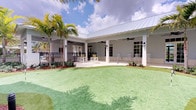 This single-story property showcases a classic Florida coastal architectural style, characterized by a standing-seam metal roof, clean grey exterior walls, and large multi-pane windows and French doors that invite abundant natural light into the interior. The expansive covered outdoor patio serves as a central gathering space, supported by white square columns and equipped with ceiling fans to ensure comfortable airflow year-round. The patio is furnished with bar-height seating in a bright yellow and white palette, as well as classic white rocking chairs arranged along the perimeter, creating a relaxed and welcoming atmosphere. A wrought-iron railing borders one side of the patio, adding a refined architectural detail. Beyond the covered area, a well-maintained artificial putting green stretches across the grounds, offering a recreational amenity set against a backdrop of mature palm trees and carefully curated tropical landscaping with vibrant mulched garden beds. The overall ambiance is bright, airy, and resort-like, blending indoor comfort with outdoor leisure in a design well-suited to a warm, sunny climate.