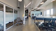 This contemporary single-floor office space offers a thoughtfully designed work environment that balances open collaboration with private focus areas. The layout features a central corridor with polished concrete floors that connects distinct functional zones throughout the space.

The open-plan work area accommodates multiple workstations arranged along long communal tables with acoustic dividers, providing a degree of privacy within a shared environment. Large windows line the perimeter, flooding the workspace with abundant natural light and offering views of the surrounding neighborhood. Soaring ceilings with exposed ductwork and linear pendant lighting add an industrial character to the interior.

Private offices and focused work pods are enclosed by floor-to-ceiling glass partition walls, maintaining visual transparency while offering acoustic separation. Modular panel dividers further define semi-private workstations within the open floor plan.

A welcoming lounge area near the entrance features comfortable seating, including rocking chairs and accent pieces, along with a sliding barn door that adds a warm, eclectic touch. A chalkboard wall serves as a creative focal point along the main corridor.

The fully equipped kitchen is a standout amenity, featuring granite countertops, wood cabinetry, a large commercial refrigerator, stainless steel appliances, and bold yellow accent walls — a vibrant contrast to the cooler tones found throughout the rest of the office.