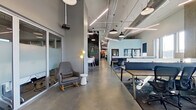 This contemporary single-floor office space offers a thoughtfully designed work environment that balances open collaboration with private focus areas. The layout features a central corridor with polished concrete floors that connects distinct functional zones throughout the space.

The open-plan work area accommodates multiple workstations arranged along long communal tables with acoustic dividers, providing a degree of privacy within a shared environment. Large windows line the perimeter, flooding the workspace with abundant natural light and offering views of the surrounding neighborhood. Soaring ceilings with exposed ductwork and linear pendant lighting add an industrial character to the interior.

Private offices and focused work pods are enclosed by floor-to-ceiling glass partition walls, maintaining visual transparency while offering acoustic separation. Modular panel dividers further define semi-private workstations within the open floor plan.

A welcoming lounge area near the entrance features comfortable seating, including rocking chairs and accent pieces, along with a sliding barn door that adds a warm, eclectic touch. A chalkboard wall serves as a creative focal point along the main corridor.

The fully equipped kitchen is a standout amenity, featuring granite countertops, wood cabinetry, a large commercial refrigerator, stainless steel appliances, and bold yellow accent walls — a vibrant contrast to the cooler tones found throughout the rest of the office.