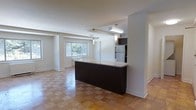 This single-floor space showcases an open-concept layout that creates a seamless flow between the kitchen and living areas. The kitchen is appointed with dark cabinetry, a stainless steel refrigerator, and a striking peninsula-style breakfast bar topped with a light stone countertop. Pendant lighting hangs above the bar, adding both function and style to the space. The warm-toned parquet flooring extends throughout the main living area, lending a classic and inviting character to the interior. Recessed and overhead lighting illuminates the space efficiently, complementing the natural light that filters in through a large window in the living area, which offers a view of lush greenery outside. The open layout, clean white walls, and thoughtful architectural details — including a stepped ceiling feature — create a bright, airy ambiance that feels both functional and comfortable. The overall design balances practicality with a welcoming aesthetic, making it well-suited for everyday living and entertaining alike.