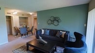 This single-floor residence showcases an open-concept design that seamlessly connects the living and dining areas, creating a cohesive and functional layout. The living room serves as the focal point of the space, anchored by a tufted dark charcoal sofa adorned with decorative blue geometric and tan accent pillows. A bold teal-green accent wall adds a striking visual element, complemented by an abstract black metal wall sculpture that enhances the modern aesthetic. A dark wood coffee table grounds the seating area, while a patterned area rug adds texture underfoot. The textured ceiling adds architectural interest throughout. Adjacent to the living area, the dining space features a round table with sleek grey chairs, offering an intimate setting for meals. Warm-toned curtains in the background introduce a subtle contrast to the cooler color palette of the living space. The flooring transitions smoothly between areas, reinforcing the open flow of the layout. Overall, the space presents a well-coordinated, contemporary interior with a balanced mix of dark furnishings, cool accent tones, and decorative details that contribute to a polished and inviting atmosphere.