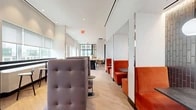 Located at 2-20 Malt Dr in Long Island City, NY, this thoughtfully designed dining space offers a versatile and welcoming environment across a single, generously proportioned floor. The interior features a clean, contemporary aesthetic defined by white walls, light wood flooring, and high ceilings that lend an open, airy feel throughout.

The layout accommodates a variety of seating configurations to suit different needs. A row of semi-private booths upholstered in rich burnt-orange velvet lines one side, each tucked between white partial-height partitions and accented by a textured tile accent wall and a globe pendant light. Opposite, tall tufted gray lounge chairs offer a more relaxed seating option. Communal long wooden tables with warm yellow stools provide collaborative or casual dining areas, while smaller round tables with bentwood and molded chairs are arranged along the window-lined perimeter.

Natural light flows in generously through large windows, framing views of the Long Island City skyline. A hallway connects to a separate dining alcove enclosed by dramatic steel-framed glass partition walls, housing an intimate table for small group gatherings. Brass railing accents and recessed lighting add refined touches throughout, creating a polished yet approachable atmosphere well-suited for dining, socializing, or focused work.