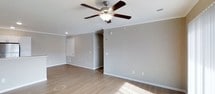 This single-floor space presents a well-maintained and straightforward layout designed for comfortable everyday living. The main living area is finished with light hardwood-style flooring that runs throughout, lending warmth and continuity to the space. Neutral gray-toned walls provide a versatile backdrop, complemented by crisp white crown molding that adds a subtle touch of architectural detail along the ceiling perimeter. A classic five-blade ceiling fan with an integrated light fixture is centrally positioned, ensuring both air circulation and ambient lighting. Recessed lighting further supplements the natural light that streams in, casting bright, even illumination across the open floor. White baseboards frame the room cleanly, and multiple electrical outlets are conveniently distributed along the walls. A visible access panel and HVAC vent indicate practical utility features built into the space. A doorway leads to an additional area, suggesting further rooms beyond the main living space. Overall, the space conveys a clean, move-in ready atmosphere with functional finishes and a neutral palette well-suited to a variety of furnishing styles and personal tastes.