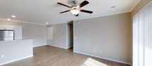 This single-floor space presents a well-maintained and straightforward layout designed for comfortable everyday living. The main living area is finished with light hardwood-style flooring that runs throughout, lending warmth and continuity to the space. Neutral gray-toned walls provide a versatile backdrop, complemented by crisp white crown molding that adds a subtle touch of architectural detail along the ceiling perimeter. A classic five-blade ceiling fan with an integrated light fixture is centrally positioned, ensuring both air circulation and ambient lighting. Recessed lighting further supplements the natural light that streams in, casting bright, even illumination across the open floor. White baseboards frame the room cleanly, and multiple electrical outlets are conveniently distributed along the walls. A visible access panel and HVAC vent indicate practical utility features built into the space. A doorway leads to an additional area, suggesting further rooms beyond the main living space. Overall, the space conveys a clean, move-in ready atmosphere with functional finishes and a neutral palette well-suited to a variety of furnishing styles and personal tastes.