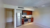 This single-floor space centers around a compact yet functional kitchen that serves as its defining feature. The kitchen is outfitted with a full suite of stainless steel appliances, including a refrigerator, gas range, built-in microwave, and dishwasher, providing a modern and cohesive aesthetic. Rich, warm-toned wood cabinetry lines the upper wall, offering ample storage, while granite countertops add a touch of refinement to the workspace. A stainless steel sink with a contemporary faucet is integrated into the counter, completing the practical layout. The kitchen flows naturally into an open adjacent area, creating a sense of connected, flexible living space. Light-toned tile flooring runs throughout, contributing to a clean and neutral foundation. A textured ceiling adds subtle visual character, and a track lighting fixture over the kitchen provides targeted, adjustable illumination. A ceiling-mounted HVAC vent ensures climate control is efficiently managed. Neutral wall tones throughout keep the space feeling bright and adaptable, while an entry door with a peephole is visible, indicating a thoughtfully organized layout from entryway to kitchen.