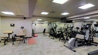 This fitness center offers a comprehensive workout environment designed to accommodate a variety of exercise routines. The space is outfitted with a solid selection of cardio equipment, including stationary bikes, elliptical trainers, and a recumbent bike, providing ample options for cardiovascular training. A multi-station cable machine anchors the strength training area, complemented by a weight bench, resistance accessories, and a stability ball for functional fitness exercises. A exercise mat on the floor adds a dedicated area for stretching or floor-based workouts. The interior features a drop ceiling with recessed panel lighting that ensures the space is well-lit throughout. Large mirrors line one wall, enhancing the sense of space and allowing users to monitor their form during workouts. A wall-mounted television is positioned for easy viewing from most areas of the room. The flooring consists of neutral-toned carpet tiles, providing comfort underfoot while maintaining a clean, professional aesthetic. The overall layout is open and thoughtfully arranged, allowing for easy movement between equipment stations. The space presents a functional and practical fitness facility suited for a range of fitness levels and training goals.