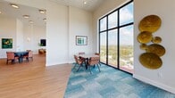 Located on an upper floor of a modern building in Miami, FL, this thoughtfully designed amenity space offers a generous open layout spanning two interconnected areas. The space is defined by dramatic floor-to-ceiling window walls on multiple sides, flooding the interior with abundant natural light and providing sweeping panoramic views of the Miami skyline and surrounding cityscape.

The main gathering area features a contemporary lounge furnished with plush teal accent chairs, a camel-toned sofa, and a patterned accent chair, all arranged atop a teal modular carpet. A slatted white ceiling adds architectural texture overhead, while bold geometric wall art contributes a modern aesthetic. This area transitions seamlessly into a dedicated game and activity room, where multiple game tables — including chess and card tables — are arranged on warm wood-look flooring. Tan leather chairs surround each table, offering comfortable seating throughout.

A smaller breakout or meeting nook is set apart from the main area, featuring its own table and seating set against another expansive glass window wall with elevated city views. Throughout the space, recessed lighting, wall sconces, and framed artwork complete the polished, upscale atmosphere, making this a versatile venue well-suited for social gatherings, leisure, and recreational activities.