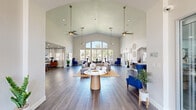 This single-story community clubhouse offers a generously sized, open-concept layout designed to impress and accommodate. The centerpiece is a dramatic great room with soaring vaulted ceilings that amplify the sense of openness and grandeur. Expansive arched windows flood the space with natural light and frame lush outdoor views, creating a seamless connection between interior and exterior. Rich wood-tone flooring runs throughout, providing a warm, cohesive foundation beneath the bright white walls and contemporary furnishings. The main lounge area features ample seating arrangements with tufted sofas, bold cobalt blue accent chairs, and glass-topped coffee tables, all anchored by a natural fiber area rug. Ceiling fans with pendant lighting hang from the vaulted ceiling, adding both style and function. A dedicated leasing office, accessible through full-height glass doors, provides a polished professional environment with individual workstations and arched architectural details. A welcoming entry foyer with a gilded ceiling accent and French doors invites natural light from the exterior. Additional utility is provided by a clean kitchenette area with white cabinetry, gold hardware, and a stainless steel sink. Decorative wall accents, vibrant indoor plants, and thoughtfully curated art complete the refined, modern ambiance of this well-appointed community space.