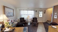 This two-floor property offers a well-organized layout that blends professional and residential functionality. The ground floor opens into a welcoming reception area furnished with classic tufted leather wingback chairs, a glass side table, and sheer curtains framing a front-facing window. A front entry door with decorative wreath detail adds a warm, approachable character. Adjacent to the reception area is a dedicated workspace featuring a long wooden desk, bulletin board, glass-top display table, and framed landscape artwork, creating a functional yet comfortable office environment.

The kitchen is outfitted with oak cabinetry, a dual-basin sink, dishwasher, and ample counter space. Parquet-style flooring adds warmth underfoot, while vertical blinds along a sliding glass door bring in natural light and offer views of the surrounding greenery. A nearby dining area accommodates a table with seating and benefits from the same outdoor view.

The upper floor hosts multiple bedrooms, each carpeted and naturally lit through standard windows. The bedrooms are comfortably scaled, featuring textured ceilings, and are accompanied by wood furniture including dressers, nightstands, and wardrobes. The overall ambiance throughout the property is understated and practical, with a neutral palette that creates a calm, functional atmosphere across both levels.