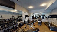 Located at 201 Orchard Hills Dr in Jeffersonville, Indiana, this well-equipped fitness center offers a comprehensive workout environment spread across a single level. The space is divided into two distinct zones, each designed to support a variety of exercise routines.

The cardio and free weights area features multiple treadmills, an elliptical, a stationary bike, and a stair climber, complemented by a full rack of dumbbells and a medicine ball for functional training. Large wall-mounted mirrors amplify the sense of space and allow for form checks during workouts. A wall-mounted television adds entertainment during cardio sessions. The flooring in this zone consists of durable, textured carpet tile suited for high-traffic use.

The strength and stretching zone offers a multi-function cable machine, a weight bench, and a dedicated wall-mounted TRX suspension training system complete with instructional guides. This area is finished with neutral carpet and benefits from abundant natural light streaming through large French doors. Those doors open directly to an outdoor pool area, providing a seamless connection between the indoor fitness space and outdoor amenities. The light-toned walls and flush ceiling fixtures throughout give the facility a clean, functional aesthetic conducive to focused training.