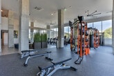 Located near Centennial Olympic Park in the heart of Atlanta, GA, this contemporary fitness center is designed to deliver a comprehensive workout experience within a spacious, well-appointed single-floor layout. Floor-to-ceiling windows line the perimeter, flooding the interior with abundant natural light and offering views of the surrounding Atlanta skyline and greenery.

The cardio zone is equipped with a row of treadmills, ellipticals, and stationary bikes, all positioned to take advantage of the panoramic city views. Adjacent to this area, a dedicated strength training section features a full rack of dumbbells, adjustable benches, and wall-mounted mirrors. A multi-functional training rig with cable systems, TRX suspension trainers, and a heavy bag anchors the strength and functional training zone, supported by rubber flooring throughout.

A distinctive open area with a bold hexagonal-patterned floor in blue and white tones provides ample space for stretching, group classes, or bodyweight training, accented by exposed concrete columns that contribute to the industrial-modern aesthetic.

The lounge area transitions seamlessly from the fitness floor, featuring wood-look flooring, blue upholstered seating, a wall-mounted display, and vibrant artwork, creating an inviting space to relax. Tall perforated wall panels and soaring ceilings complete the refined, energetic atmosphere of this facility.