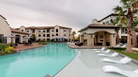 This amenity space combines an expansive fitness center with a resort-style outdoor pool area, creating a comprehensive wellness destination. The fitness center is housed within an architecturally distinctive building featuring soaring vaulted ceilings, pendant globe lighting, and a row of grand arched windows that flood the interior with natural light while offering views of the surrounding landscaping and pool area. The gym floor is generously proportioned, accommodating a full lineup of cardio machines — including treadmills, ellipticals, stair climbers, and stationary bikes — along one side, while the opposite side hosts a robust strength training section with cable machines, free weights, medicine balls, battle ropes, and TRX suspension equipment. Full-length mirrors amplify the sense of space throughout. Adjacent to the main gym floor is a dedicated studio room, separated by decorative white lattice-panel barn doors, featuring built-in cubbies and access to a spin or yoga room with frosted glass doors. Outside, the expansive freeform swimming pool is complemented by a stone-accented covered pavilion with an outdoor fireplace, mounted TV, and seating, along with additional poolside lounge chairs and canopied sun areas.