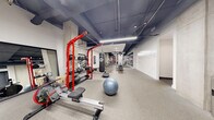 This expansive fitness center is designed to accommodate a wide range of training disciplines within a single, thoughtfully organized floor. The space features a modern industrial aesthetic, with exposed dark ceilings, visible ductwork and structural columns, and clean white walls that create a striking visual contrast. Linear LED lighting runs throughout, providing bright, even illumination across all workout zones.

The facility is divided into several distinct training areas. A dedicated cardio zone houses treadmills and stationary cycling bikes, complemented by a wall-mounted television for entertainment during workouts. An adjacent open floor area with wood-style flooring provides space for stretching, rowing machines, and functional training activities.

The strength training section is comprehensively outfitted with a multi-station cable machine, a selection of selectorized weight machines, and a bold red power rack complete with a full dumbbell rack and medicine balls. Rubber flooring throughout this zone provides durability and safety. A boxing area features a heavy punching bag suspended from the red rack frame, alongside a balance disc and stability ball for functional conditioning.

Full-length mirrors are positioned strategically throughout the space to assist with form and technique. The facility also includes accessible restroom facilities and a water fountain, ensuring a complete and user-friendly workout environment.