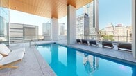 Located at 900 Rue Saint-Jacques in the heart of downtown Montréal, this expansive rooftop terrace offers a sophisticated outdoor amenity space with sweeping panoramic views of the city skyline. The terrace is finished with large-format stone tiles throughout and is organized into distinct functional zones that together create a cohesive, resort-inspired environment.

At the heart of the terrace sits a semi-covered lap pool, sheltered beneath a striking warm-toned wood-paneled ceiling supported by substantial concrete columns. Lounge chairs are arranged poolside, and the open-air setting allows unobstructed views of Montréal's iconic downtown towers on all sides.

Surrounding the pool, the open terrace provides generously proportioned gathering areas furnished with a mix of round bistro tables, bar-height communal tables, and modern seating arrangements. Deep navy planters filled with ornamental grasses serve as elegant dividers between zones, adding a layer of greenery to the urban setting.

A glass-curtain-wall pavilion anchors one end of the terrace, its floor-to-ceiling glazing and wood ceiling continuing the refined architectural language of the covered pool area. Bronze-toned framing details add warmth to the otherwise crisp, contemporary palette of concrete, glass, and steel. Black metal railings define the perimeter, ensuring safety while preserving the open, airy character of this elevated urban retreat.