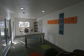 The Village Oaks game room is a single-floor recreational amenity space designed to foster community interaction and entertainment. The room is finished with bright white walls and patterned carpet tiles featuring colorful hexagonal accents in teal, yellow, orange, and gray, creating a modern and energetic atmosphere. A centerpiece white foosball table anchors the main area, while an octagonal poker table with padded surface and built-in cup holders provides a dedicated space for card games. A wall-mounted flat-screen television is positioned nearby for additional entertainment. A high-top bistro table with bar-height chairs sits along the windowed wall, allowing natural light to filter in and providing views of the surrounding greenery. A black metal shelving unit offers storage for games and accessories, and a restroom is conveniently accessible within the space. Large glass doors connect the game room to an adjacent lounge area, which is visible through interior windows and features a fireplace, colorful seating, and bold patterned flooring. Oversized Scrabble-tile wall art spelling "Village Oaks" adds a playful, branded touch to the décor. The overall design is clean, contemporary, and welcoming, making it an ideal gathering spot for socializing and recreation.