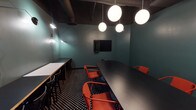 Located on W 8th Place in the heart of Los Angeles, CA, this single-floor meeting space delivers a striking blend of modern design and functional layout. The room is defined by deep teal walls that create an immersive, focused atmosphere, complemented by a dark, exposed ceiling with industrial-style ductwork that adds an urban edge.

The flooring is a bold black-and-white polka-dot pattern that grounds the space with graphic visual energy. At the center sits a large, curved matte black conference table surrounded by vibrant red-orange chairs, offering ample seating for collaborative meetings or presentations. Along one wall, a secondary counter-height work surface with light wood cabinetry provides additional workspace for attendees.

Lighting is a standout design element, featuring a cluster of spherical globe pendant lights that cast a warm, even glow throughout the room. Articulating arm wall sconces add supplementary task lighting and a sculptural quality to the teal walls. A wall-mounted flat-screen display is positioned for easy viewing from all seating areas, making the space well-equipped for presentations and video conferencing. Glass-paneled walls offer a view into the adjacent corridor, maintaining a sense of openness while preserving the room's dedicated, professional character.