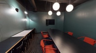 Located on W 8th Place in the heart of Los Angeles, CA, this single-floor meeting space delivers a striking blend of modern design and functional layout. The room is defined by deep teal walls that create an immersive, focused atmosphere, complemented by a dark, exposed ceiling with industrial-style ductwork that adds an urban edge.

The flooring is a bold black-and-white polka-dot pattern that grounds the space with graphic visual energy. At the center sits a large, curved matte black conference table surrounded by vibrant red-orange chairs, offering ample seating for collaborative meetings or presentations. Along one wall, a secondary counter-height work surface with light wood cabinetry provides additional workspace for attendees.

Lighting is a standout design element, featuring a cluster of spherical globe pendant lights that cast a warm, even glow throughout the room. Articulating arm wall sconces add supplementary task lighting and a sculptural quality to the teal walls. A wall-mounted flat-screen display is positioned for easy viewing from all seating areas, making the space well-equipped for presentations and video conferencing. Glass-paneled walls offer a view into the adjacent corridor, maintaining a sense of openness while preserving the room's dedicated, professional character.