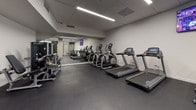 This fitness center offers a comprehensive workout environment designed to accommodate a wide range of exercise needs. The space is finished with dark rubber flooring throughout, providing both durability and traction, while a dropped ceiling with flush linear LED lighting ensures consistent, even illumination across the entire floor.

The cardio section lines one wall and includes multiple treadmills and elliptical trainers, each equipped with digital display screens for performance tracking. Adjacent to this zone, a dedicated strength training area houses a full dumbbell rack with an extensive weight range, a Matrix cable machine, a Smith machine with an adjustable bench, and additional selectorized weight equipment. Spin bikes round out the cardiovascular options for low-impact training.

Full-length mirrors extend along key walls, visually expanding the space and allowing users to monitor their form. A wall-mounted television adds an entertainment element to the environment. Frosted glass partitions and a structural concrete column add architectural character, while the neutral gray-and-white palette contributes to a clean, professional atmosphere. The overall layout is thoughtfully organized to allow multiple users to train simultaneously without congestion.