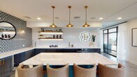 Located at 921 W First St in Winston-Salem, NC, this single-floor space delivers a polished, amenity-rich environment defined by thoughtful design and a cohesive blend of modern and industrial aesthetics. The layout flows across several distinct zones, each with its own character while maintaining a unified visual language throughout.

The kitchen area is a standout feature, showcasing navy cabinetry with brass hardware, a luminous white subway tile backsplash, and illuminated open shelving. A large white quartz island anchors the space, surrounded by upholstered bar seating in warm neutral tones. Three brass pendant lights hang overhead, and a decorative branded medallion adds a distinctive focal point to the design.

Adjacent lounge areas offer varied seating arrangements suited to both social and relaxed settings. One zone features a rich cognac leather sofa paired with neutral accent chairs, set against exposed brick columns and floor-to-ceiling windows that draw in natural light and frame outdoor views. A vertical wood slat partition adds architectural warmth and defines the space.

A media lounge highlights a bold feature wall of diagonal wood slats set against teal-patterned wallcovering, housing a mounted flat-screen display. Elsewhere, a quieter seating nook with cobalt blue accent chairs sits before large windows overlooking an outdoor terrace, with polished concrete flooring and geometric tile transitions enhancing the contemporary feel.