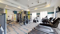 This fitness center is a thoughtfully designed workout facility that combines functionality with modern aesthetics. The space features tray ceilings with recessed lighting and ceiling fans, creating a comfortable and well-lit environment for exercise. Bold teal and chartreuse accent stripes along the walls add a vibrant, energetic atmosphere, complementing the neutral tones of the patterned carpet flooring.

The facility is comprehensively equipped for both cardio and strength training. A row of Matrix treadmills lines one wall, positioned alongside large windows fitted with roller shades that allow natural light to filter in while maintaining privacy. Elliptical machines and stationary bikes provide additional cardio options, while a central cable machine tower and a variety of selectorized strength equipment serve those focused on resistance training. Free weights and adjustable benches round out the strength training area.

Multiple wall-mounted flat-screen TVs are positioned throughout the space, ensuring entertainment is accessible from virtually any piece of equipment. Full-length mirrors along one wall visually expand the room and allow users to monitor their form. Access is provided through glass-paned double doors, offering a welcoming entry to the space. The overall layout is open and spacious, allowing for comfortable movement between equipment stations.