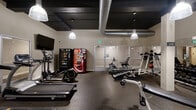 The fitness center at 211 Warren Ave in Baltimore, Maryland, is a well-equipped, single-floor facility designed to support a comprehensive workout experience. The space features an industrial-inspired aesthetic, with a dark-painted ceiling that showcases exposed silver ductwork and pendant lighting fixtures that cast a warm, focused glow throughout the room.

The cardio section includes multiple SportsArt treadmills, an elliptical trainer, a rowing machine, and a recumbent stationary bike, providing a range of low- and high-impact options. A wall-mounted flat-screen television is positioned near the cardio equipment for entertainment during workouts. The strength training area is equipped with a multi-station cable machine, a dedicated free weights rack stocked with medicine balls and dumbbells, and additional benches and resistance equipment.

Large mirrored wall panels visually expand the space and allow users to monitor their form during exercise. Durable rubber flooring covers the entire area, offering safety and impact absorption. Snack and beverage vending machines are conveniently located near the entrance, adding a practical amenity for users. The space is accessed through glass-paneled double doors and maintains a clean, functional layout suited for a variety of fitness routines.