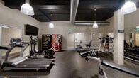 The fitness center at 211 Warren Ave in Baltimore, Maryland, is a well-equipped, single-floor facility designed to support a comprehensive workout experience. The space features an industrial-inspired aesthetic, with a dark-painted ceiling that showcases exposed silver ductwork and pendant lighting fixtures that cast a warm, focused glow throughout the room.

The cardio section includes multiple SportsArt treadmills, an elliptical trainer, a rowing machine, and a recumbent stationary bike, providing a range of low- and high-impact options. A wall-mounted flat-screen television is positioned near the cardio equipment for entertainment during workouts. The strength training area is equipped with a multi-station cable machine, a dedicated free weights rack stocked with medicine balls and dumbbells, and additional benches and resistance equipment.

Large mirrored wall panels visually expand the space and allow users to monitor their form during exercise. Durable rubber flooring covers the entire area, offering safety and impact absorption. Snack and beverage vending machines are conveniently located near the entrance, adding a practical amenity for users. The space is accessed through glass-paneled double doors and maintains a clean, functional layout suited for a variety of fitness routines.