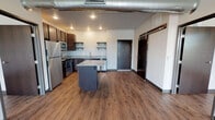 This single-floor unit offers a thoughtfully designed open-concept layout with a clean, modern aesthetic throughout. The kitchen serves as a central feature, equipped with stainless steel appliances including a refrigerator, range, microwave, and dishwasher. Rich dark cabinetry is complemented by light quartz-style countertops, open floating shelves, and a central island that provides additional workspace and storage. A glass backsplash adds a sleek finishing touch.

Warm hardwood-style flooring runs consistently throughout the entire unit, creating a cohesive and inviting atmosphere. The two bedrooms are generously sized, each featuring ceiling fans, large windows that invite natural light, and direct access to walk-in closets with built-in shelving. The open layout of each bedroom allows for flexible furniture arrangements.

The bathroom is well-appointed with a navy blue vanity, framed mirror, a soaking tub, and matching hardwood-style flooring that ties the space together. Exposed ductwork visible in the main living area introduces an industrial design element, adding character and contrast to the otherwise clean, neutral interior. Dark-framed interior doors and bold black baseboards provide sharp, modern detailing throughout the unit.