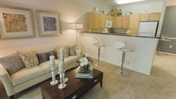 This single-floor residence offers an open-concept layout that seamlessly connects the living area and kitchen, creating a cohesive and functional living space. The living area is carpeted in neutral tones and features a comfortable sofa accented with decorative pillows, a dark wood coffee table with drawer storage, and elegant silver candle holders as centerpiece décor. A floor lamp provides warm ambient lighting, complementing the framed wall art that adds a refined touch to the space.

The kitchen is positioned directly adjacent to the living area, separated by a raised breakfast bar with dark countertops and fitted with two sleek, modern bar stools. Light maple cabinetry lines the upper walls, offering generous storage, and extends to the top of the cabinetry where decorative accessories are displayed. The kitchen is equipped with built-in appliances, including a microwave, and features a functional countertop workspace. The open bar-style connection between the kitchen and living room enhances the overall sense of space, making the unit well-suited for both everyday living and casual entertaining.