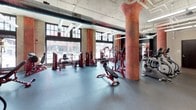 This fitness facility masterfully blends industrial architecture with a fully outfitted workout environment. The space is defined by its soaring exposed ceilings with visible ductwork and structural systems, large multi-pane windows that open onto street views and invite abundant natural light, and striking weathered columns that serve as dramatic visual anchors throughout the floor.

The main workout area is expansive and thoughtfully organized, featuring a comprehensive selection of red-accented strength machines, free weights, cable systems, and power racks arranged across durable rubber flooring. A large display screen is mounted prominently in the training zone, supporting guided workout programming. A dedicated cardio section houses treadmills and additional equipment, positioned near mirrored walls that enhance the sense of openness.

A welcoming entryway with warm hardwood flooring and tufted leather seating provides a comfortable transition into the facility. Restrooms and a water station are conveniently accessible from the main training floor. Throughout the space, the combination of raw brick, oxidized columns, and modern fitness equipment creates a dynamic, energizing atmosphere well-suited for focused, high-performance training.