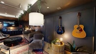 Located at 975 Pandora Ave in Victoria, BC, this single-floor amenity space offers a thoughtfully curated environment that blends music culture with sophisticated lounge design. The interior living room is the centerpiece, featuring deep charcoal walls adorned with framed vinyl records and music memorabilia, complemented by plush tufted sofas, layered Persian rugs, and an eclectic mix of vintage armchairs. Hanging greenery cascades from the ceiling tray, softening the moody, intimate atmosphere. Musical instruments — including acoustic and electric guitars and a keyboard — are displayed throughout, reinforcing the space's artistic character. Rich burgundy velvet curtains and dramatic accent lighting add warmth and depth. A glass-paneled garage-style door connects the lounge to the outdoor covered patio, which features stone tile paving, decorative floral-printed panels, and space for casual games. The exterior courtyard is a well-landscaped urban retreat, with raised planters, young trees, bold checkerboard paving, and a vibrant mural visible nearby. The building entrance is framed by a sleek modern facade of glass and masonry, with manicured hedges and planters completing the polished streetscape presentation.